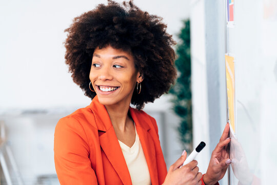 Happy businesswoman with Afro hairstyle standing by whiteboard in office