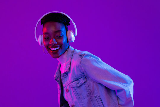 Trendy happy smiling African-American woman wearing headphones and listening to music in modern purple studio background