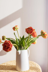 Orange red tulip flowers with green stems and leaves in a white ceramic pitcher on a light shadow white wall and window background. Creative floral botany wallpaper. Minimal creative greeting card.