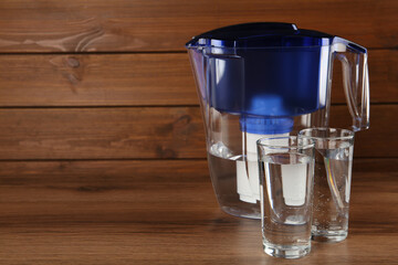 Filter jug and glasses with purified water on wooden table. Space for text