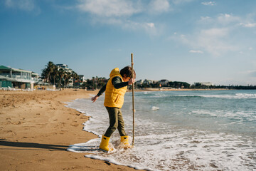 Boy in yellow rubber boots playing with stick and sand at the beach. School kid touching water at...