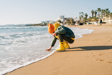 Obraz premium Boy in yellow rubber boots playing with water and sand at the beach. School kid touching water at autumn winter sea. Child having fun with waves at the shore. Spring Holiday vacation concept.