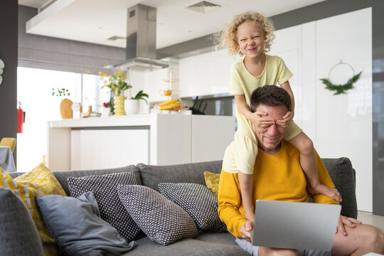 Mischievous daughter covering eyes of father sitting with laptop on sofa at home