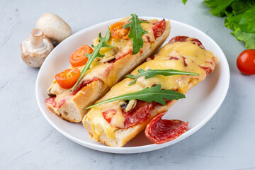 Two slices of pizza on a baguette, pizza sandwich with cheese, pepperoni sausages and mushrooms in a white round plate, gray background.