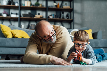 Senior man looking at grandson playing with toy car lying in living room