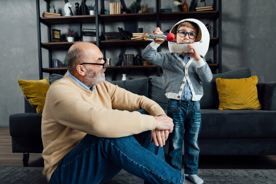 Senior man looking at grandson wearing space helmet playing with toy rocket at home