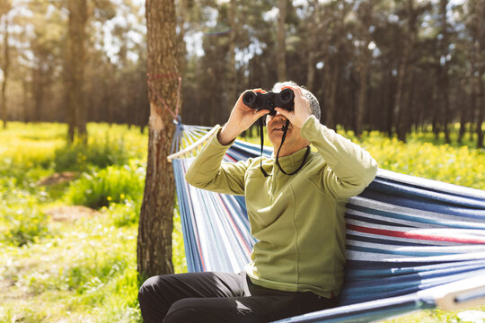 Man looking through binoculars sitting in hammock on sunny day