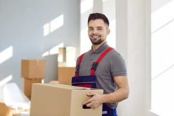 Portrait of young worker from modern professional moving company or express delivery service. Happy handsome man in workwear uniform standing inside house, holding cardboard box and smiling