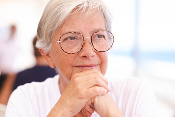 Close up portrait of a beautiful senior caucasian woman with white hair and glasses sitting outdoors while looking thoughtfully away with her hands under her chin
