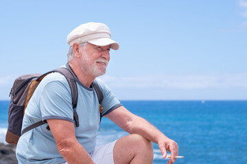 Attractive senior man with beard and hat sitting outdoors at sea looking at horizon while smoke a cigarette relaxing on a sunny day. Copy space, horizon over water. Retirement freedom vacation concept