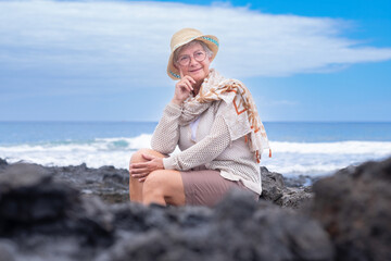 Fototapeta na wymiar Portrait of smiling senior woman wearing eyeglasses and summer hat sitting outdoors at sea enjoying good time, freedom and relax. Cloudy sky, copy space. Travel vacation retirement concept
