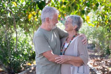 Beautiful white-haired senior couple walking in the woods hug looking in the eyes. Smiling elderly grandparents enjoy healthy lifestyle in public park
