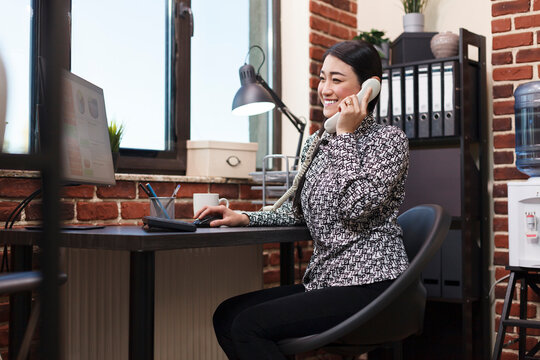 Advertising agency office smiling worker being informed on landline about correctly issued accounting documentation. Young asian manager talking on landline with team leader about promoting campaign.
