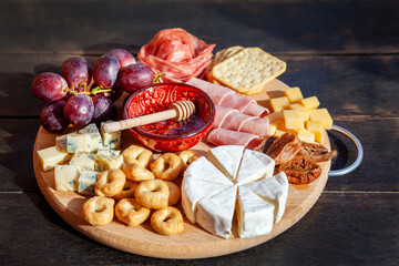Round cutting board with slicing assorted cheese, sausage rosette, ham, cracker, grapes and figs with honey