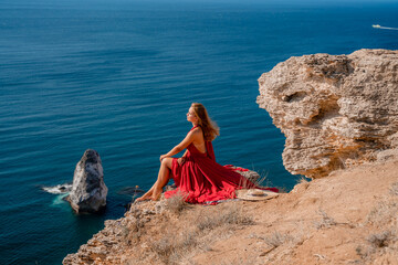 A girl with flowing hair in a long red dress sits on a rock above the sea. The stone can be seen in the sea.