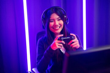 Playing video game. Young asian pretty woman sitting on chair holding joystick in living room. Happy female Professional Streamer chinese wearing headphone playing game in dark room neon light.