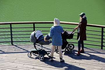 Two women walking with baby prams on lake embankment. Mothers talking in city park at spring