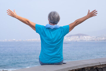 Rear view portrait of happy senior woman wearing blue tshirt and scarf enjoying outdoors freedom and vacation at sea. Nice female looking at horizon with outstretched arms