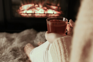 Woman with cup of tea resting near fireplace at home, closeup