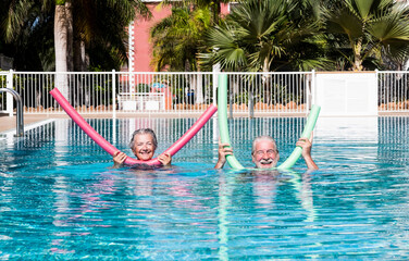 Cheerful adult happy senior couple having fun in outdoors swimming pool with swim noodles. Smiling retired people playing together in the pool water under the sun enjoying vacation