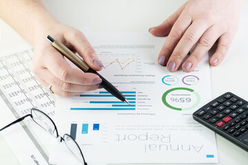 Close-up of business woman's hands holding pen and pointing at financial charts. Visible hands, pen, eyeglasses and calculator on white desk. Accounting, business, finance, tax and office concepts.