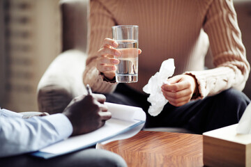 Young woman drinking water and wiping tears with napkin in therapy session with psychologist