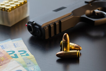 9 mm semi-automatic pistol, full metal jacket bullets and Euro banknotes laid out on black table. Conceptual mockup of gun control, crime, military-industrial complex or defense.