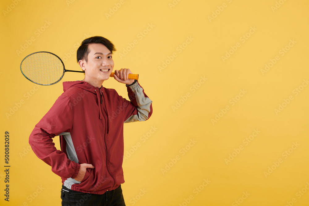 Wall mural Portrait of happy excited badminton player with racket standing against yellow background