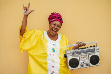 Happy senior black woman with traditional african dress dancing holding vintage stereo - Focus on...