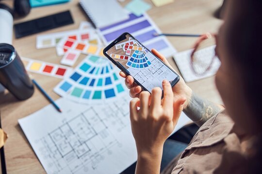 Close up shot of woman taking a photo of a blueprint and color samples palette using smartphone while working on new interior design project in her office