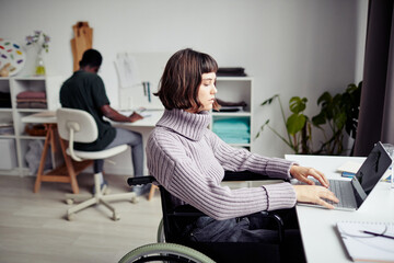 Serious young woman in wheelchair working on laptop in fashion studio with her colleague