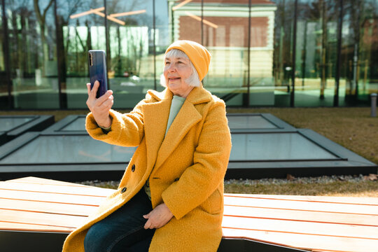 Positive smiling woman wearing casual clothes and hat holding smartphone in one hand outdoors, she sitting on a bench. Urban lifestyle concept. Traveler. Attractive redhead female chilling alone.