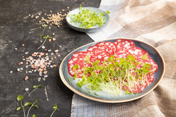 Slices of smoked cervelat salami sausage with spinach microgreen on black, side view, close up.