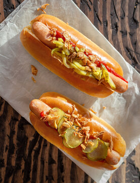 two hot dog buns with sausages pickles fried onions ketchup and mustard bottle on the table