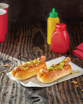 two hot dog buns with sausages pickles fried onions ketchup and mustard bottle on the table