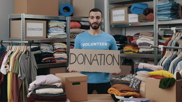 Portrait view of the caucasian volunteer man holding cupboard banner with donation word and looking at the camera with smile. Shelves with belongings at the background. Humanitarian aid concept