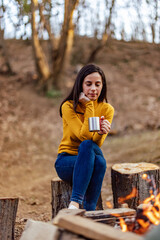The woman sitting on the log, holding a drink and it is heated by the campfire.