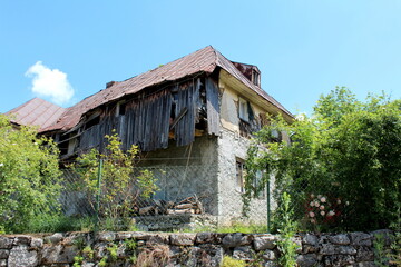 Fototapeta na wymiar Very old abandoned dilapidated cracked wooden boards suburban family house built on concrete and stone foundation covered with rusted metal roof tiles built on top of small hill surrounded with dense 