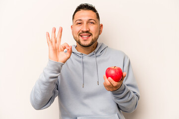 Hispanic man holding an apple isolated on white background cheerful and confident showing ok...