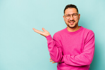 Fototapeta premium Young hispanic man isolated on blue background showing a copy space on a palm and holding another hand on waist.