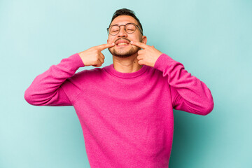 Fototapeta na wymiar Young hispanic man isolated on blue background smiles, pointing fingers at mouth.