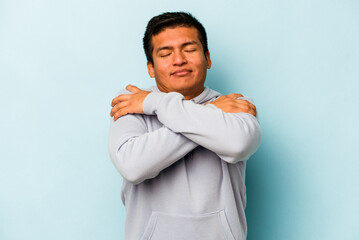 Young hispanic man isolated on blue background hugs, smiling carefree and happy.