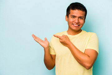 Young hispanic man isolated on blue background excited holding a copy space on palm.