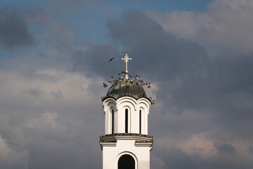 Flock of pigeons fly around church bell tower against dark stormy clouds