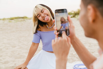 Young man taking photo of his girlfriend during picnic on beach