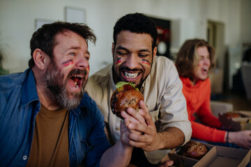 Happy German football fans friends watching football at home and eating hamburger.
