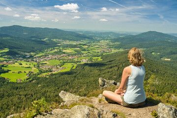 Naklejka na ściany i meble A woman sitting on mount Osser, overlooking the landscape of Lamer Winkel with the small town Lam in the Bavarian Forest