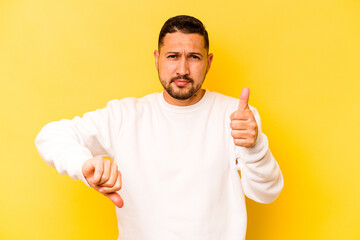 Young hispanic man isolated on yellow background showing thumbs up and thumbs down, difficult choose concept