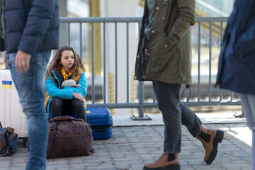 Sad Ukrainian immigrant child with luggage waiting at train station, Ukrainian war concept.