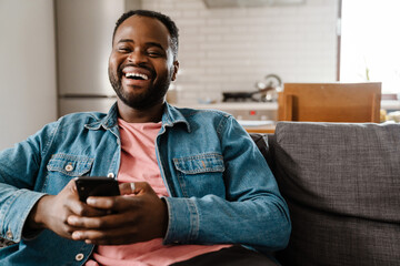 Black bristle man smiling and using cellphone while sitting on sofa